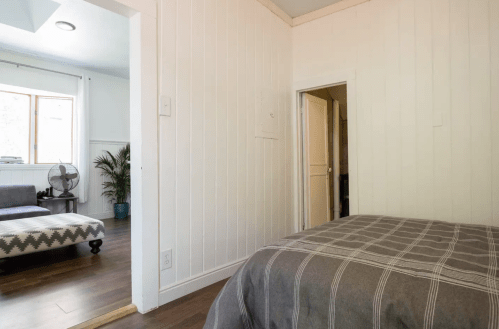 Cozy interior view of a room with a bed, wooden floor, and a doorway leading to another space with a fan and seating.