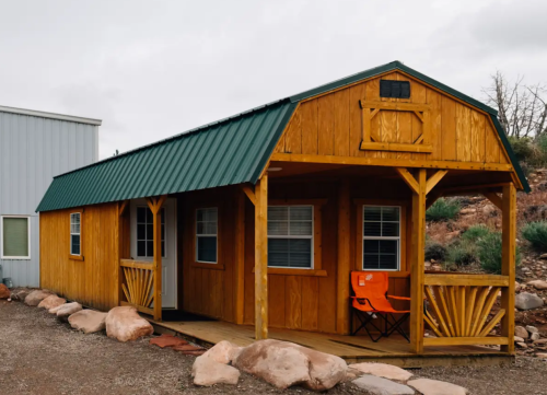 A small wooden cabin with a green metal roof, featuring a porch and an orange chair outside.