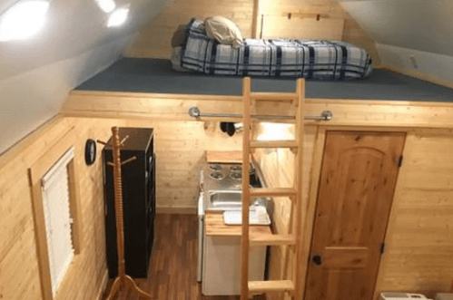 Cozy tiny home interior with a loft bed, kitchen area, and wooden accents. A ladder leads to the sleeping space.