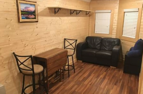Cozy wooden interior with a leather couch, small table, and two chairs, featuring a framed landscape painting on the wall.
