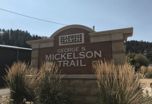 Sign for the Deadwood Trailhead, featuring the George S. Mickelson Trail, surrounded by tall grass and trees.