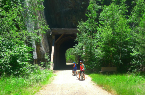 Two cyclists pause on a gravel path surrounded by lush greenery, approaching a large wooden tunnel in the distance.