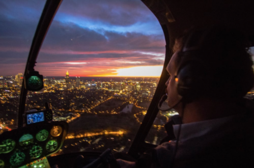 A pilot in a helicopter gazes at a vibrant sunset over a city skyline, with lights twinkling below.