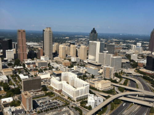 Aerial view of a city skyline featuring tall buildings, highways, and green spaces under a clear blue sky.