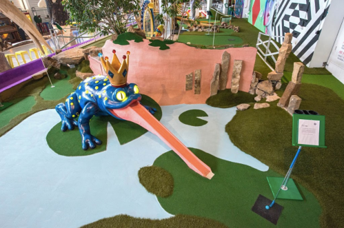 A large, colorful frog with a crown sticks out its long tongue in a whimsical mini-golf setting.