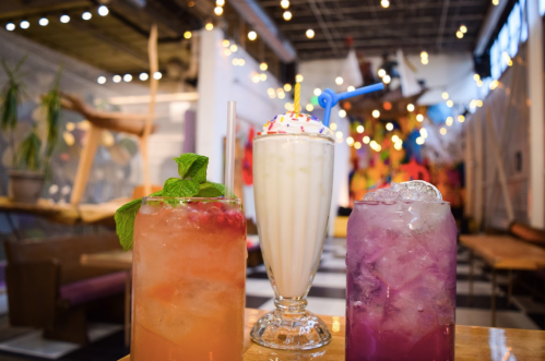 Three colorful drinks on a table: a fruity cocktail, a milkshake with sprinkles, and a purple beverage, in a vibrant setting.