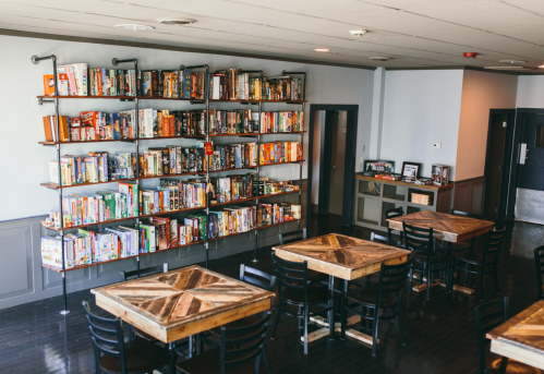 A cozy game café with wooden tables and a large shelf filled with board games.