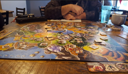 A tabletop game in progress, featuring a colorful map and various game pieces, with a player’s hand visible.