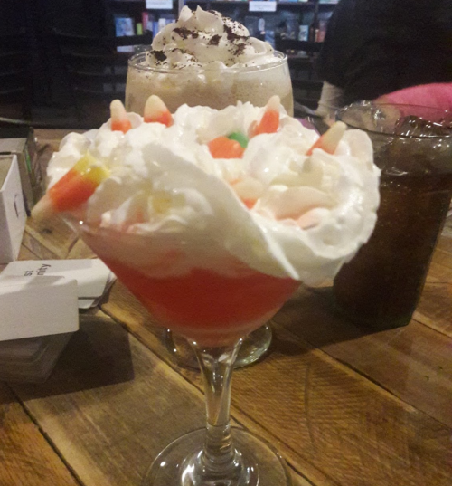 A colorful dessert in a glass topped with whipped cream and candy, with a drink in the background on a wooden table.