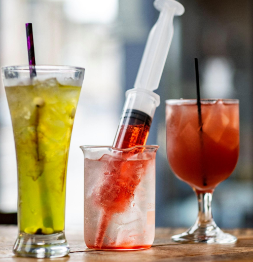 Three colorful cocktails on a wooden table: a green drink, a red drink with a syringe, and a pink drink.