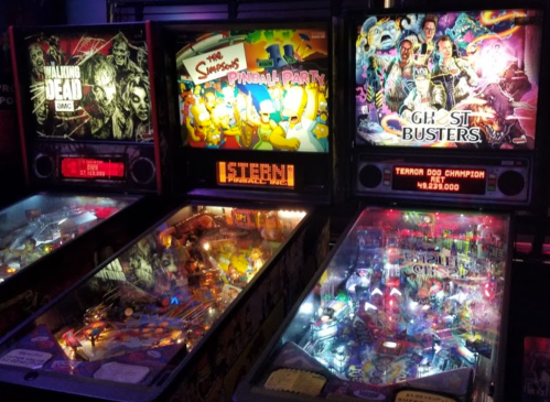 Three pinball machines featuring "The Walking Dead," "The Simpsons Pinball Party," and "Ghostbusters" in a dimly lit arcade.