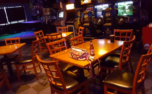 A dimly lit arcade bar with empty tables and chairs, featuring vintage arcade games in the background.