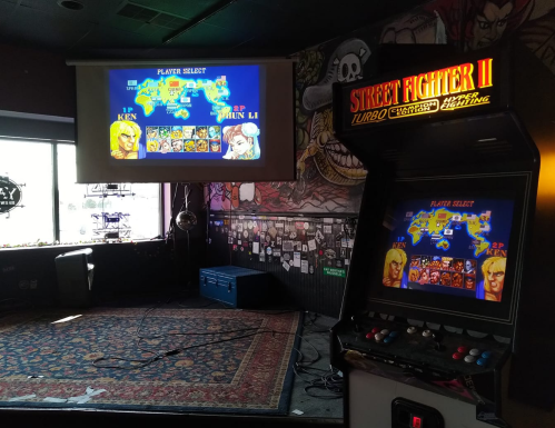 An arcade machine for Street Fighter II with a player select screen displayed on a wall in a vibrant, decorated venue.
