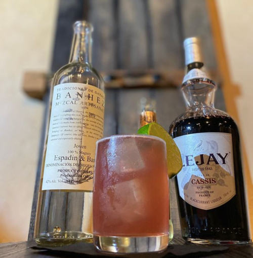 A cocktail glass with a lime wedge, next to bottles of mezcal and cassis on a wooden surface.