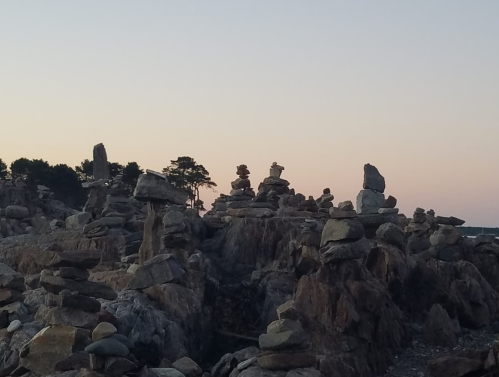 A rocky shoreline at sunset, featuring stacked stones and silhouettes of trees against a pastel sky.