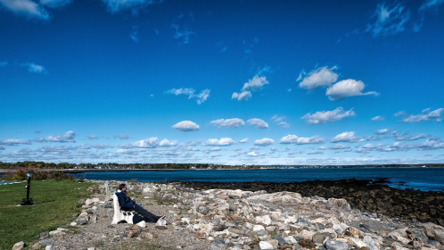 A person sits on a bench by the rocky shore, gazing at the water under a blue sky with fluffy clouds.