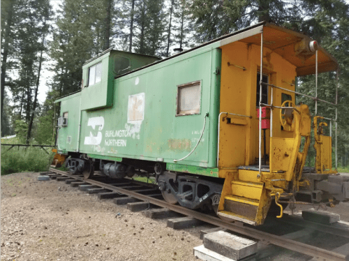 A green and yellow train caboose on tracks, surrounded by trees in a natural setting.