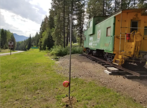 A green caboose on tracks beside a grassy area, with trees and mountains in the background. A red ball hangs nearby.