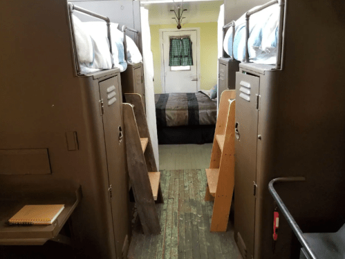 Interior view of a shared sleeping area with bunk beds, lockers, and a bedroom at the end, featuring wooden steps and a desk.