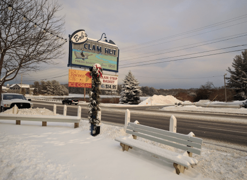 A snowy scene featuring Bob's Clam Hut sign, a decorated pole, and benches along a road in winter.