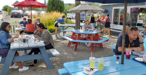Outdoor dining scene with people at colorful picnic tables, enjoying food and drinks on a sunny day.
