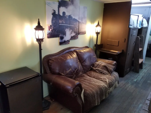 Cozy room with a brown leather couch, lamps, a train picture, and a small fridge against a green wall.