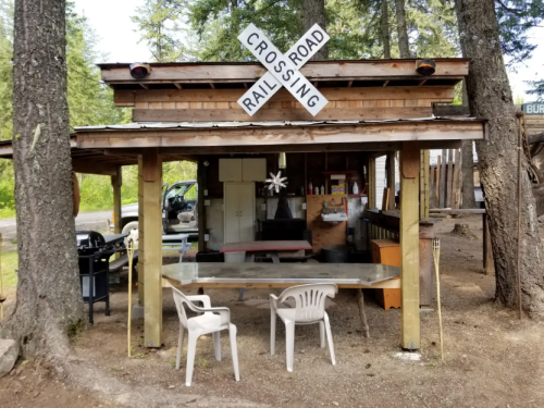 A rustic wooden structure with a railroad crossing sign, featuring a table and chairs outside in a forested area.