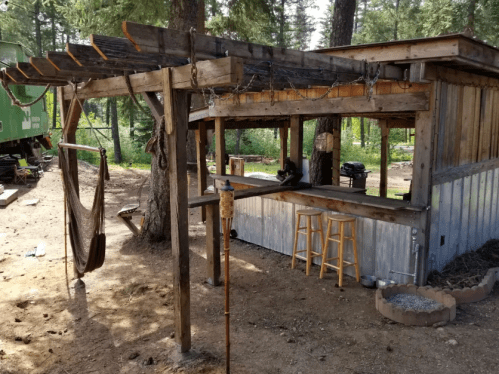 A rustic outdoor structure with a wooden bar, stools, and a hammock, surrounded by trees in a natural setting.