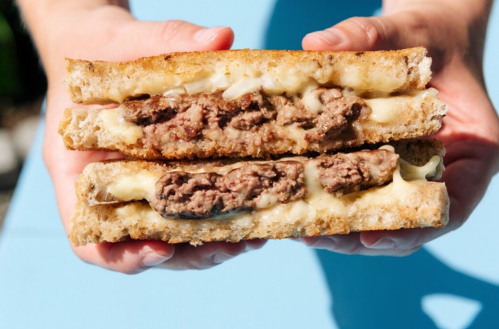 A person holds a thick sandwich with two beef patties and melted cheese between toasted bread slices.