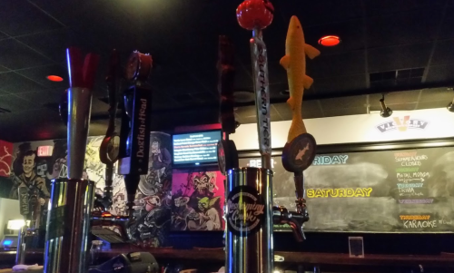 A close-up of beer taps in a bar, with colorful handles and a vibrant mural in the background.