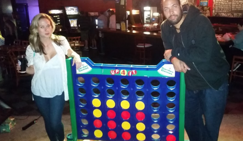 A woman and a man stand beside a large Connect Four game in a bar setting, both posing for the photo.