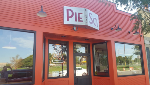 Exterior of Pie Sci pizza restaurant, featuring a red facade and a sign with a pizza slice in a beaker design.