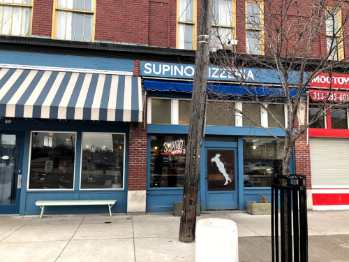 Exterior of Supino Pizzeria, featuring a blue storefront and striped awning, with a tree and utility pole nearby.