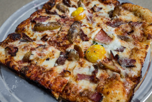 A square pizza topped with cheese, mushrooms, bacon, and two cracked eggs, served on a round tray.