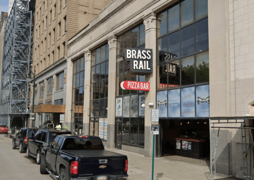 Exterior view of a building featuring "Brass Rail" and "Pizza Bar" signs, with parked cars along the street.