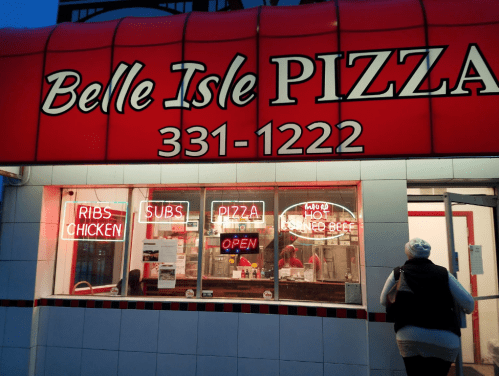 A pizza shop with a red awning and neon signs for pizza, ribs, subs, and open hours. A person is entering the shop.