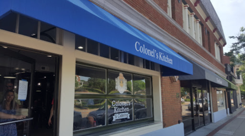 Exterior of Colonel's Kitchen with a blue awning, showcasing the restaurant's name and nearby storefronts.