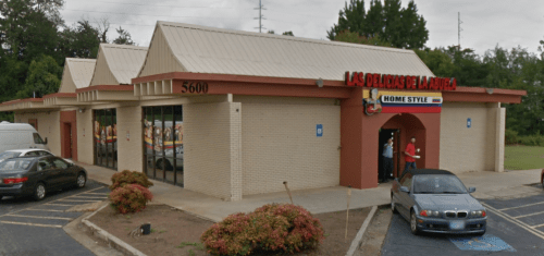 Exterior of a restaurant named "Las Delicias La Raza," featuring a sign and a parking lot with cars.