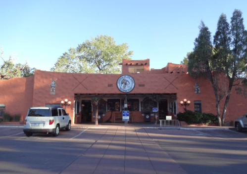 A southwestern-style building with a round sign, surrounded by trees and parked cars, under a clear blue sky.