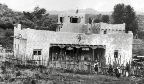Historic black-and-white photo of a southwestern-style building with a wooden fence and people in the foreground.