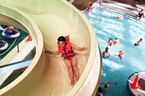 A child in a life jacket slides down a water slide into a pool filled with people enjoying the water.