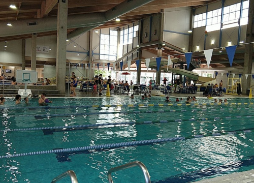 Indoor swimming pool with lanes, swimmers, and spectators. Bright windows and a water slide in the background.