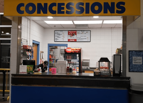 Concession stand with snacks and drinks, featuring a menu board and a staff member preparing orders.