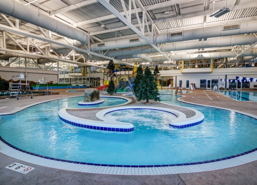 Indoor swimming pool with winding water features, trees, and a slide, surrounded by lounge areas and seating.