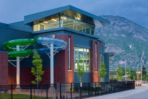 Modern building with large windows, featuring water slides, set against a mountainous backdrop under a cloudy sky.