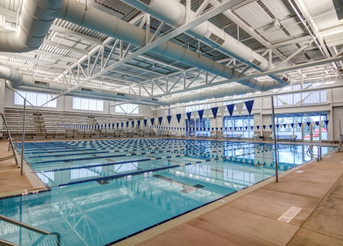 Indoor swimming pool with clear blue water, lane markings, and seating area, brightly lit with large windows.