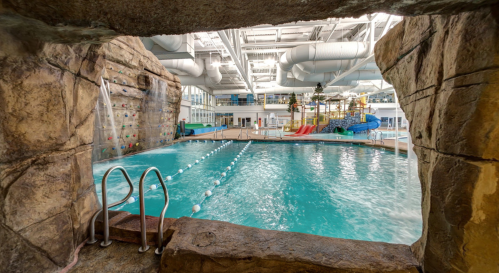 Indoor swimming pool with a rock climbing wall, water slides, and a waterfall feature, viewed through a stone archway.