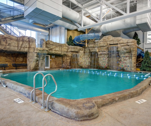 Indoor pool with rock formations, waterfalls, and a water slide, surrounded by trees and seating areas.