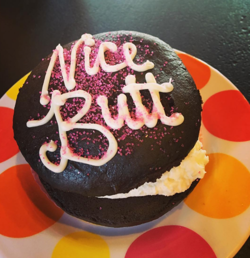 A chocolate dessert with white filling, topped with pink sprinkles and the words "Nice Butt" in icing, on a colorful plate.