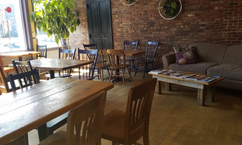 Cozy café interior with wooden tables, chairs, a couch, and a brick wall adorned with plants.
